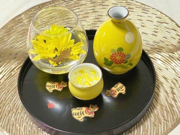 ガラスのおちょこについだ日本酒に菊を散らした菊酒と黄色の徳利が寅模様の盆に乗せられている写真。